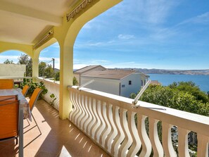 Ein Balkon mit wellenförmiger weißer Brüstung, einem Tisch mit Stühlen und Blick auf ein Gewässer und hügelige Landschaft.