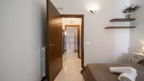 A hotel room with a bed, a radiator, a door, a hallway, and a wall-mounted shelf with a vase. - La Palma - Apartment with terrace and pool view (Lazise)