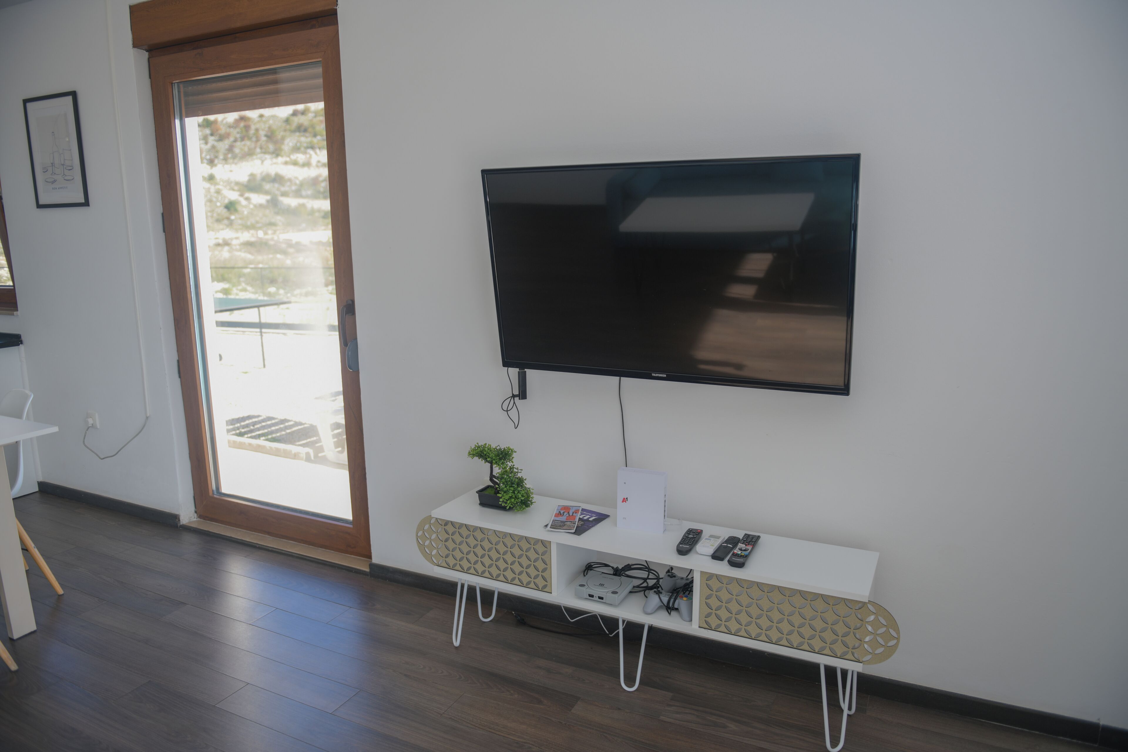 A modern living room with a flat-screen TV mounted on the wall, a white console table with a plant, and a door leading to an outdoor area.