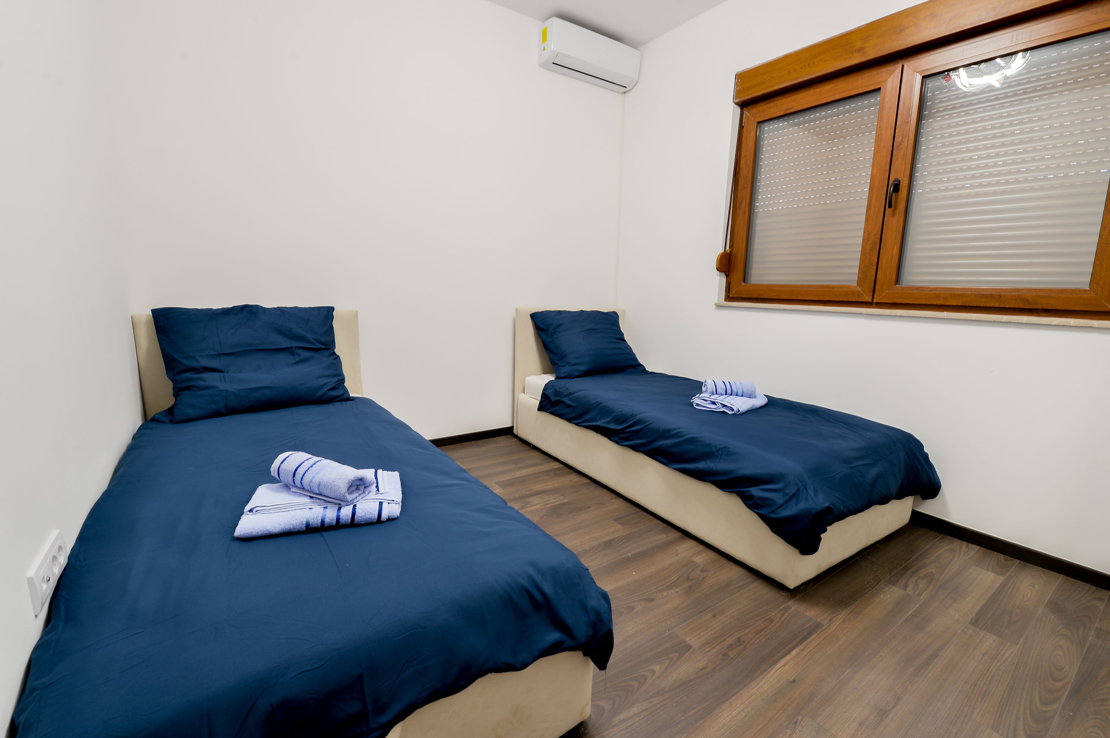 Duas camas de solteiro com roupa de cama e almofadas azul-escuras, um piso de madeira, uma janela com persianas e um aparelho de ar condicionado na parede.