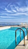 A clear blue pool with metal railings, surrounded by lounge chairs and a view of the ocean.