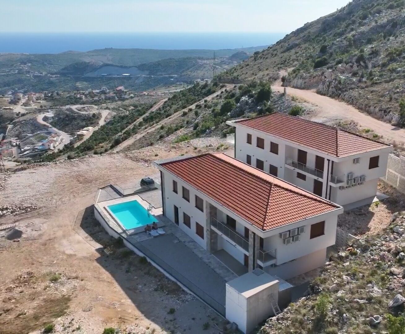 Un bâtiment moderne à deux étages, doté d’une piscine, entouré d’un paysage vallonné et offrant une vue sur la mer.
