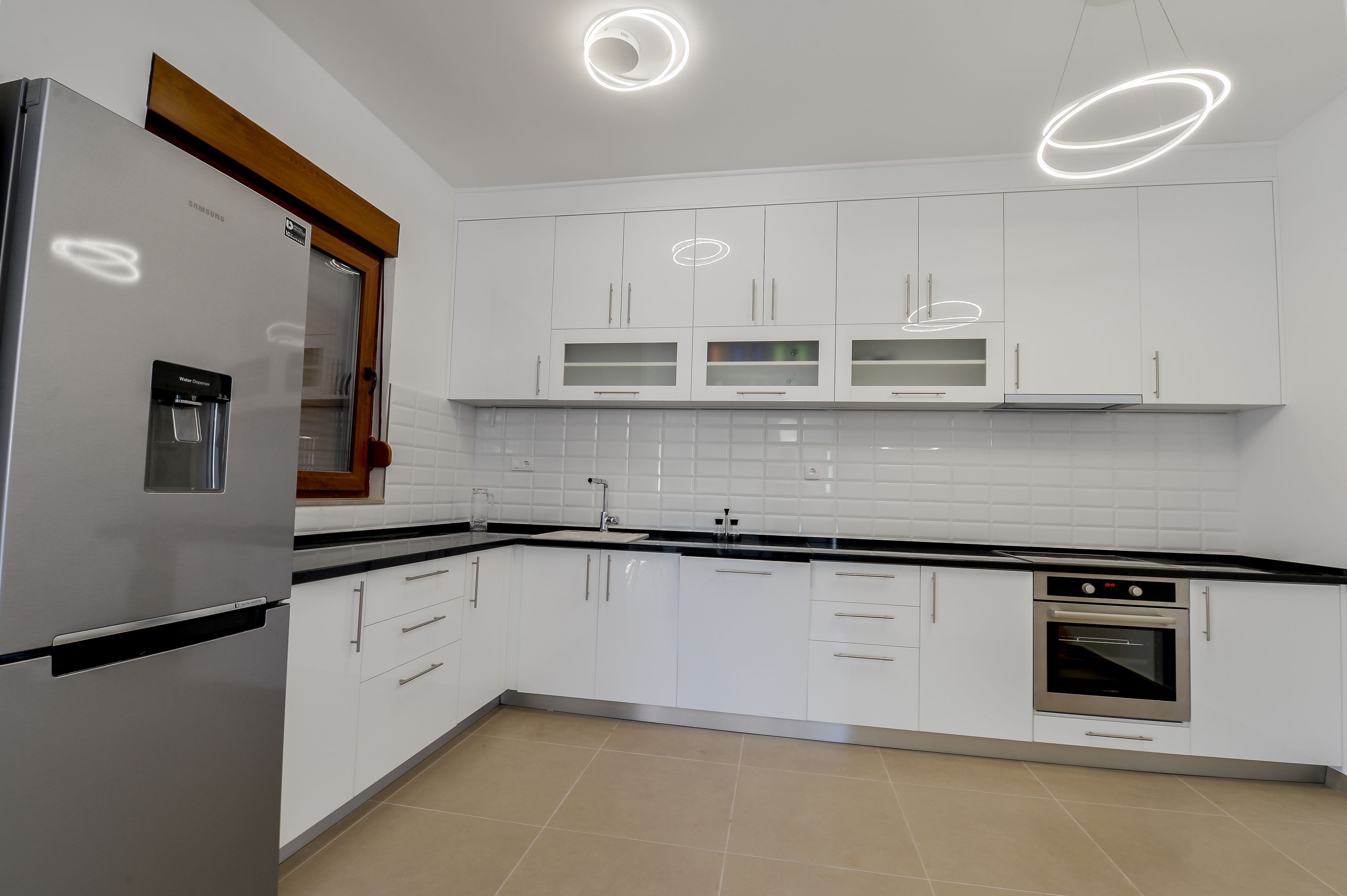 A modern kitchen with white cabinets, a stainless steel refrigerator, a built-in oven, and a water dispenser.