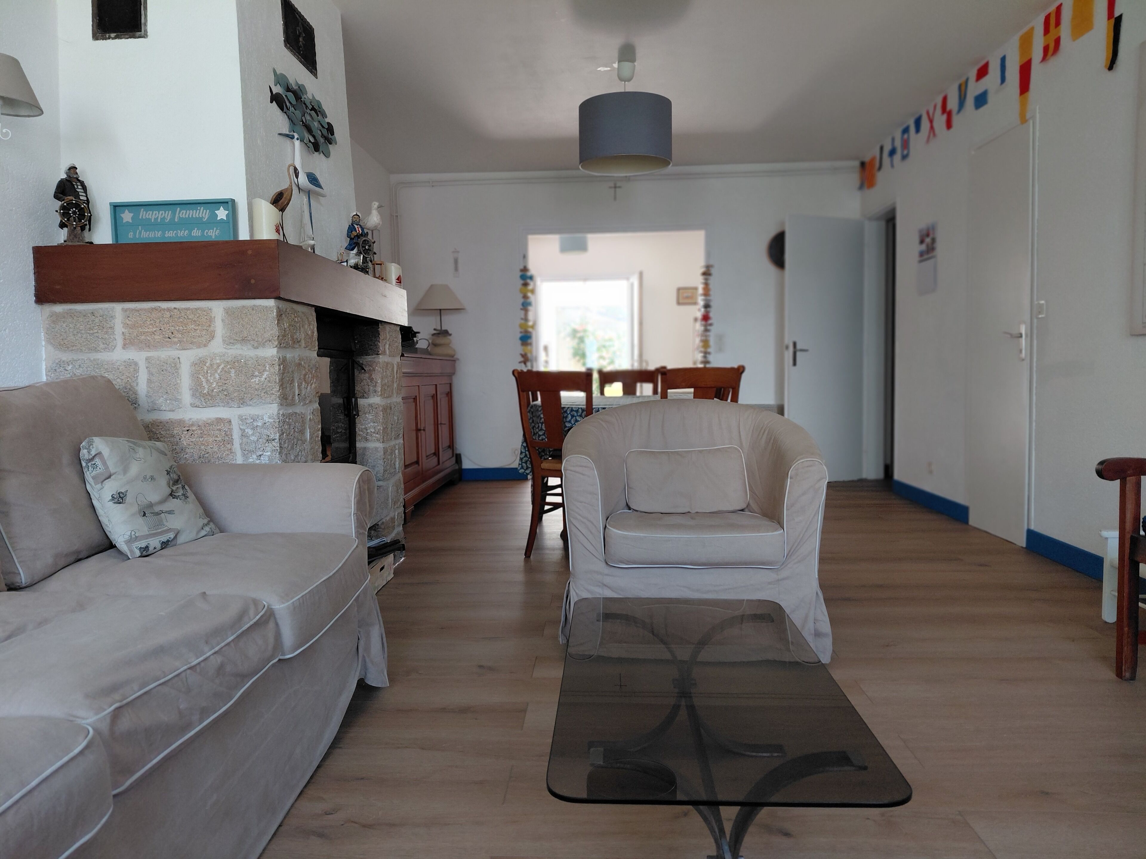 Un salon comprenant un canapé, un fauteuil et une table basse en verre. On y trouve une cheminée avec un mur en pierre, un meuble en bois et, au fond, un coin repas.