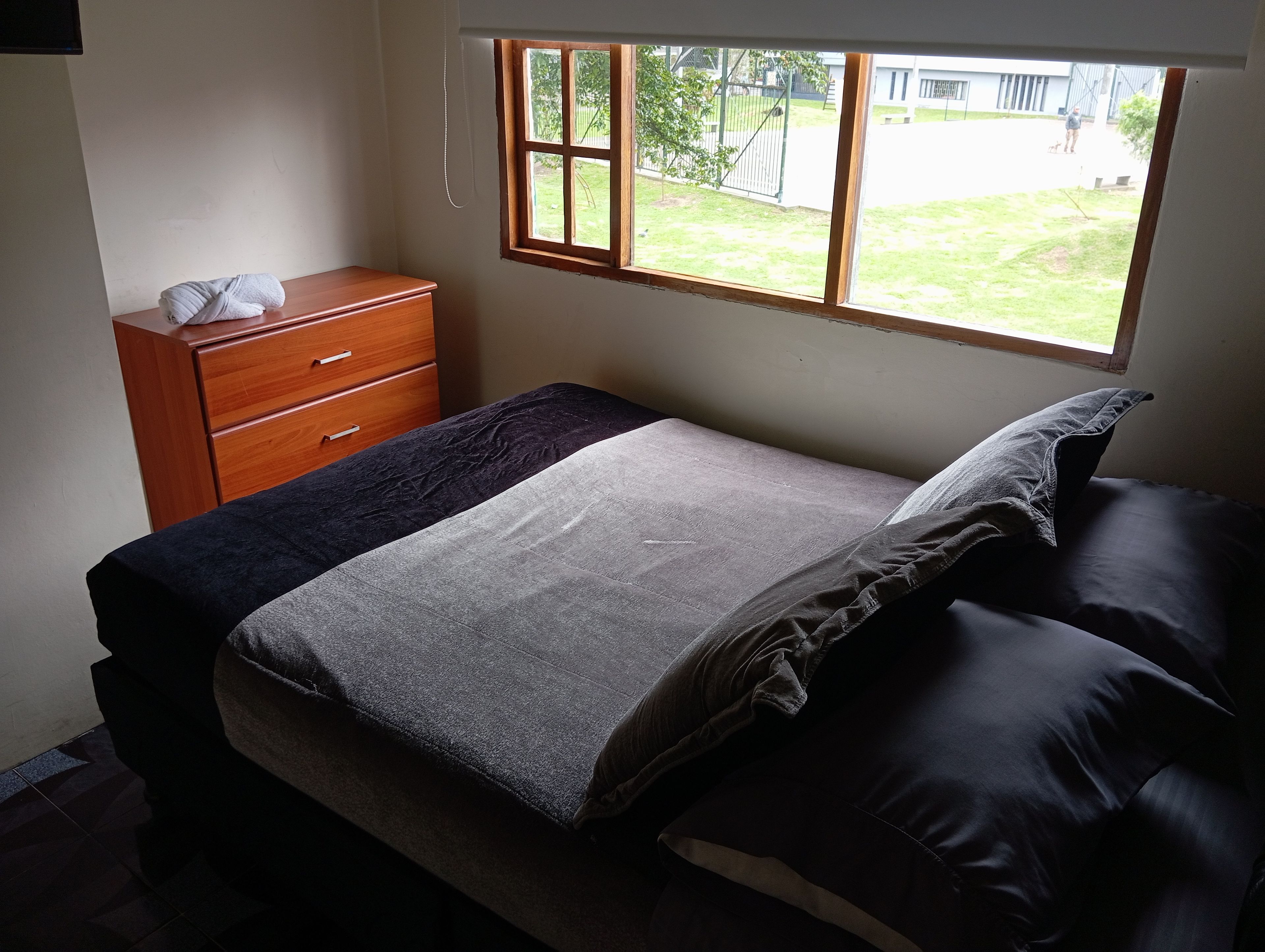 A bedroom with a bed, a wooden dresser, a window, and a view of the outside.