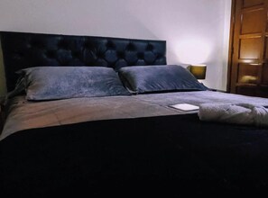 A bed with a dark headboard, two pillows, a blanket, and a bedside table with a lamp.