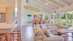 A spacious living area with a large sofa, a dining table, and a kitchen area with wooden cabinets. - Tropical & Elegant- Sleeps 8- Ocean/Waterfall Views (Princeville)