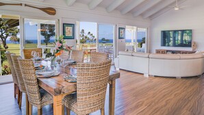 A dining area with a wooden table and wicker chairs, a white sofa, and a television in the living room. - Tropical & Elegant- Sleeps 8- Ocean/Waterfall Views (Princeville)