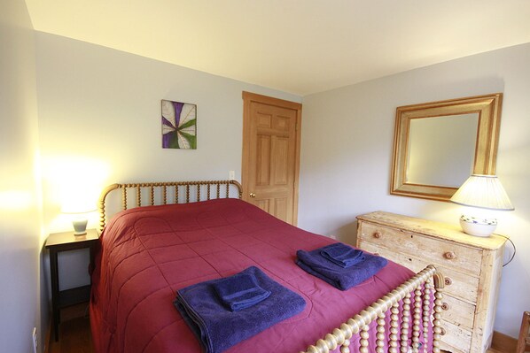 A bedroom with a wooden headboard, a red bedspread, two folded towels, a wooden dresser, a mirror, and a wall art piece.