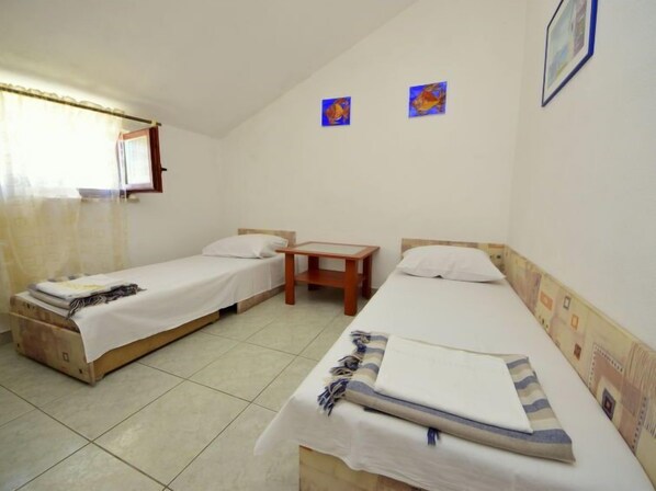 A simple room with two single beds, a small wooden table, and a window with curtains. - Margita by Interhome (Brač/Supetar)
