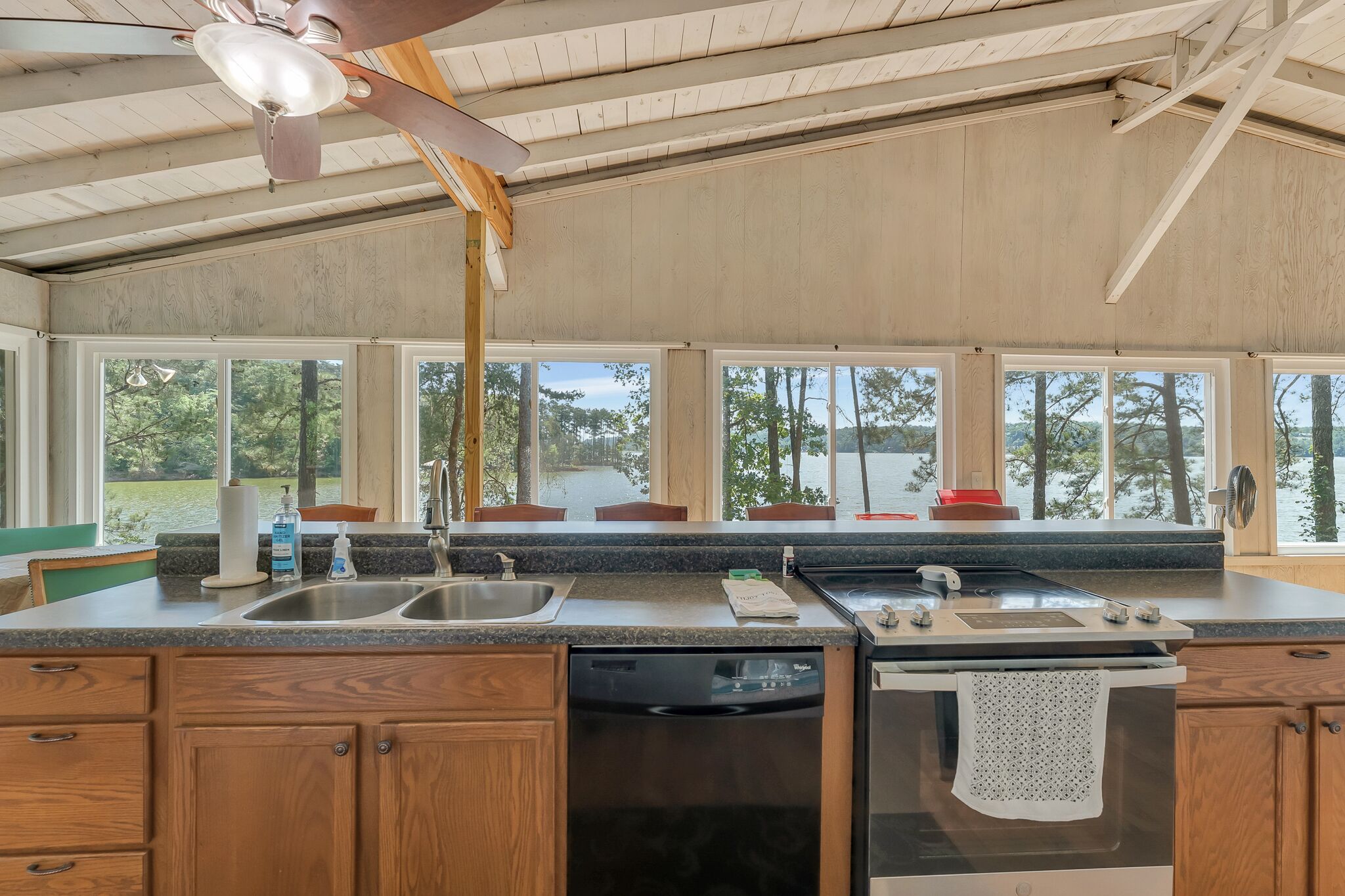 A kitchen with wooden cabinets, a dishwasher, a stove, and large windows offering a view of trees and a body of water.