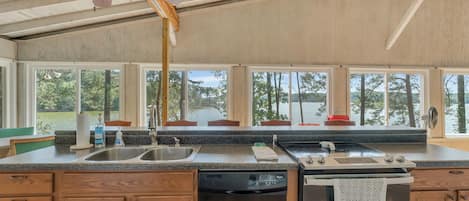 A kitchen with wooden cabinets, a dishwasher, a stove, and large windows offering a view of trees and a body of water.