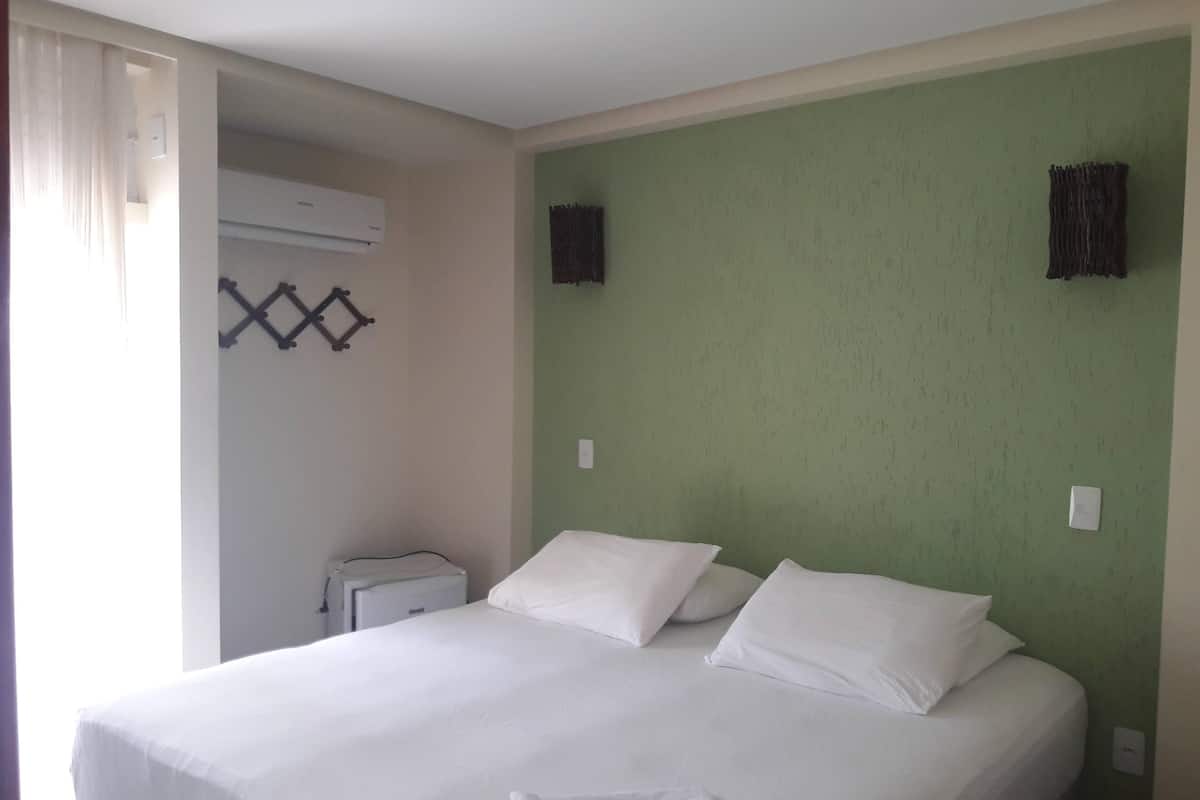 A double bed with white bedding in a room with green walls, a small bedside table, and an air conditioning unit.