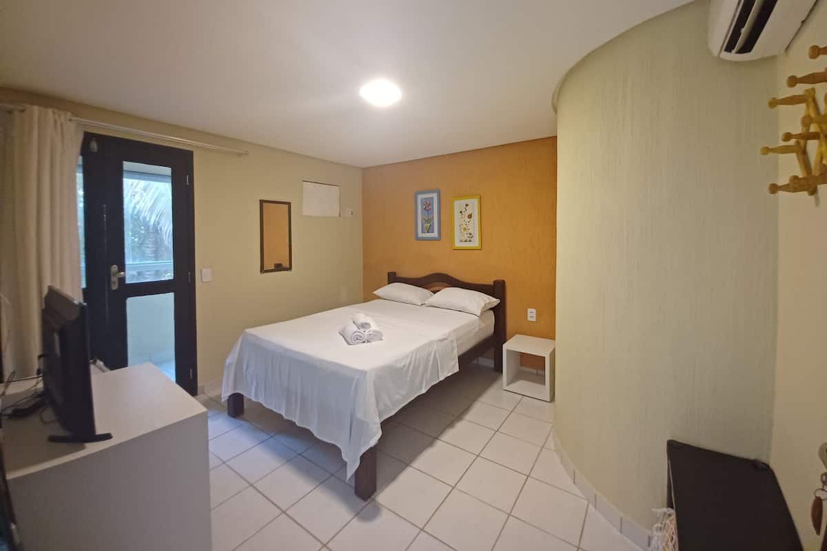 A hotel room with a bed, a desk, and a chair. There is a door with a window, a wall-mounted air conditioner, and a small bedside table.