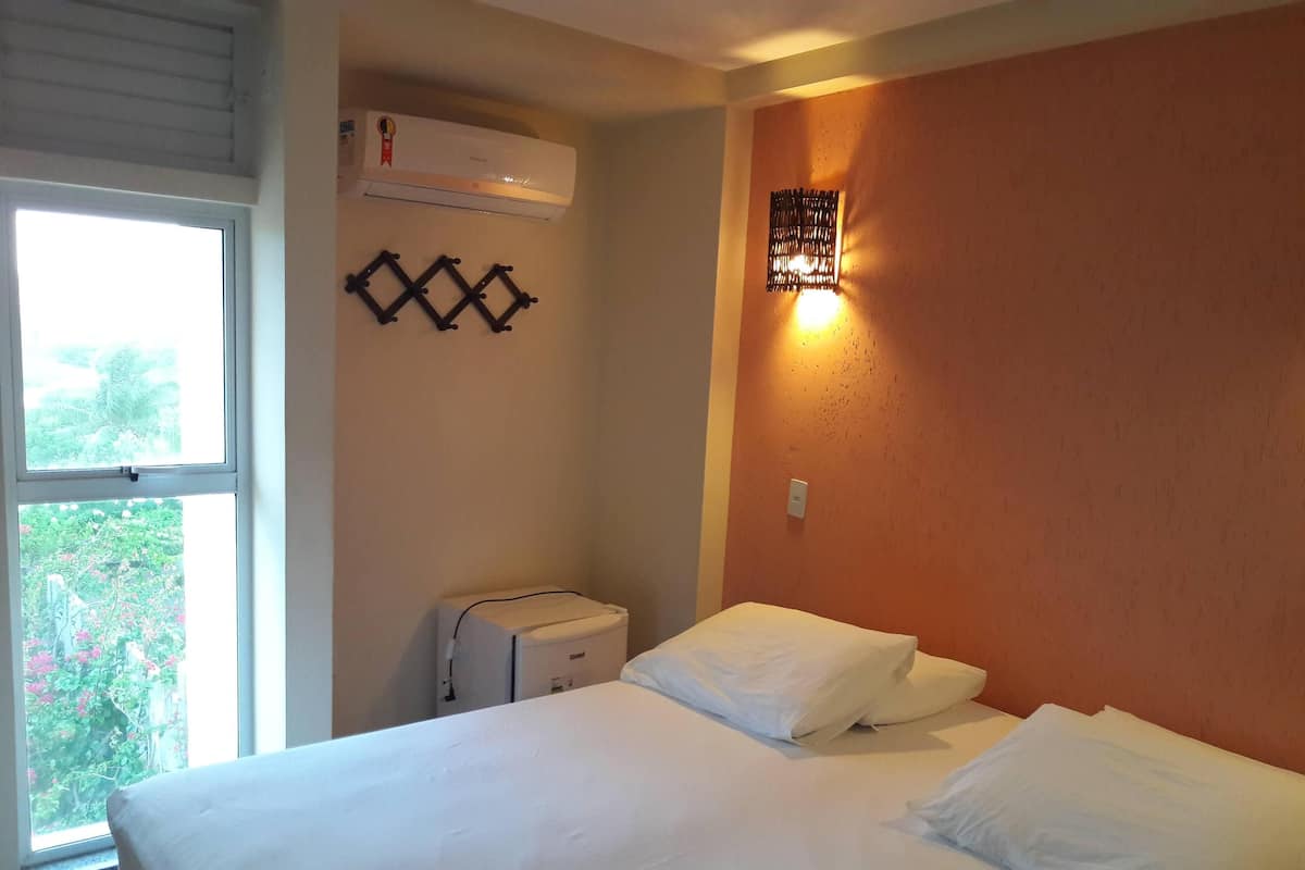 A hotel room with a bed, an air conditioner, a small table, and a window with a view of greenery.