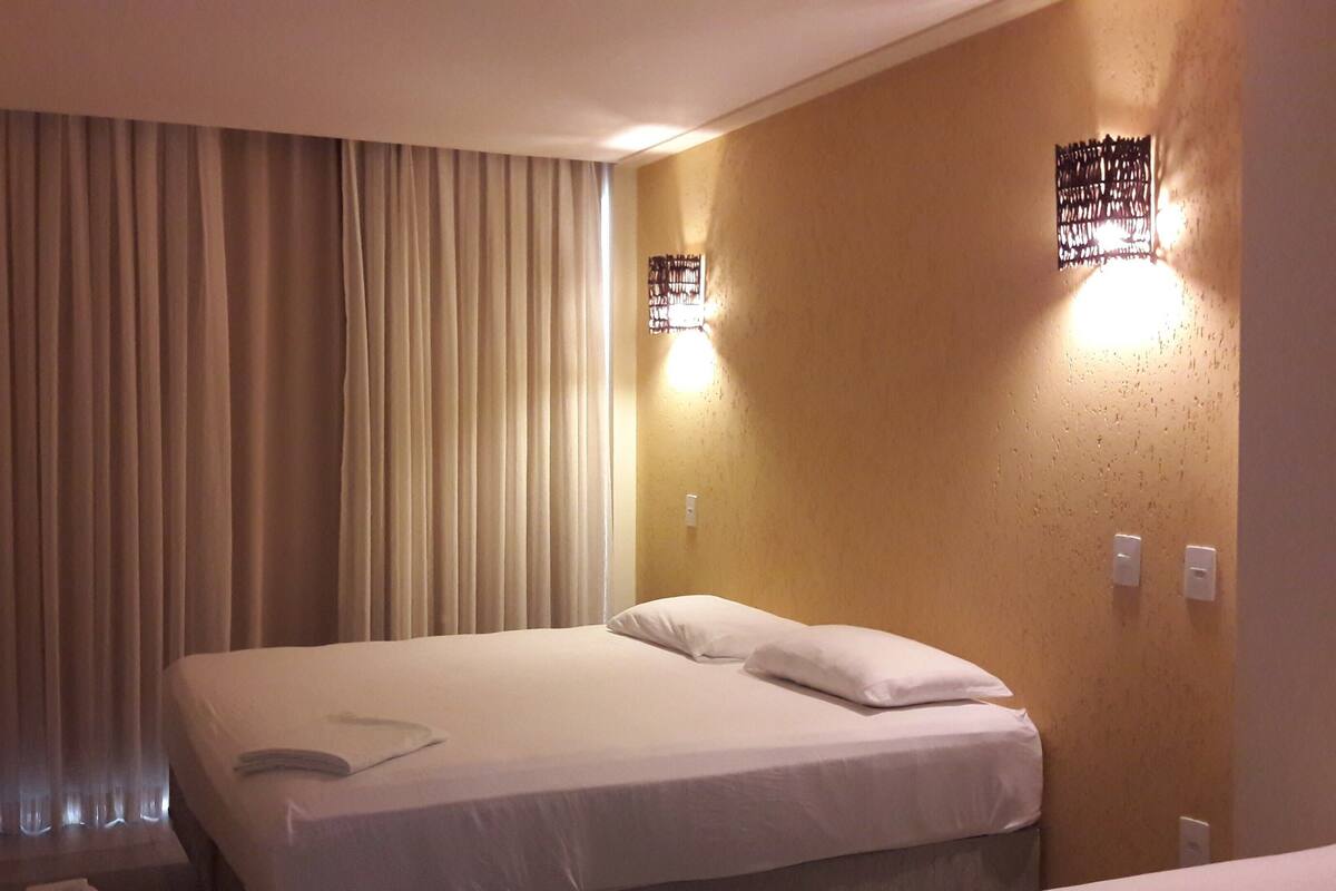 A hotel room with a bed, curtains, a wall-mounted light fixture, and a television on the wall.