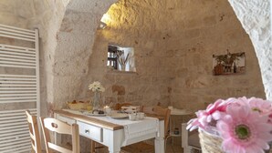 A rustic stone interior with a wooden table set for dining, a wicker chair, and a basket of flowers. - Stylish Trulli with Private Pool for 4 guests in Puglia (Castellana Grotte)