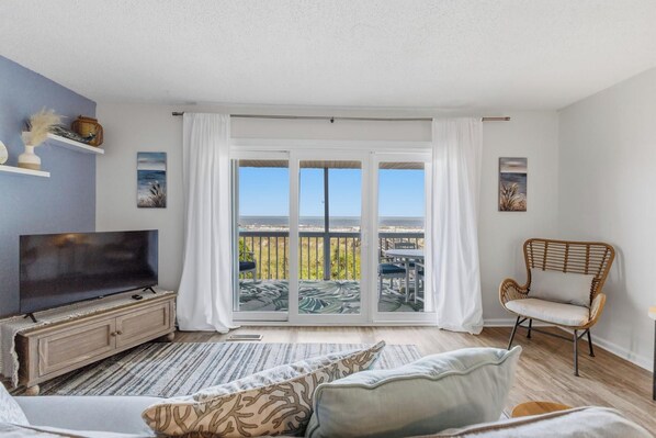 A modern living room with a television, a sofa, a chair, and a large sliding glass door leading to a balcony with a view.