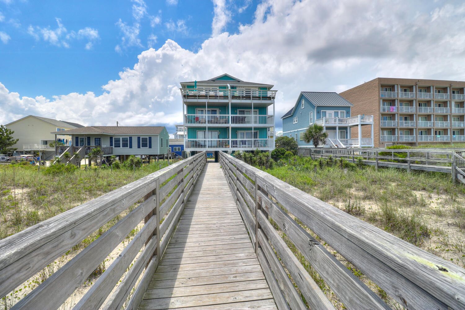 A wooden boardwalk leading to a beachfront building with multiple balconies and a blue facade.