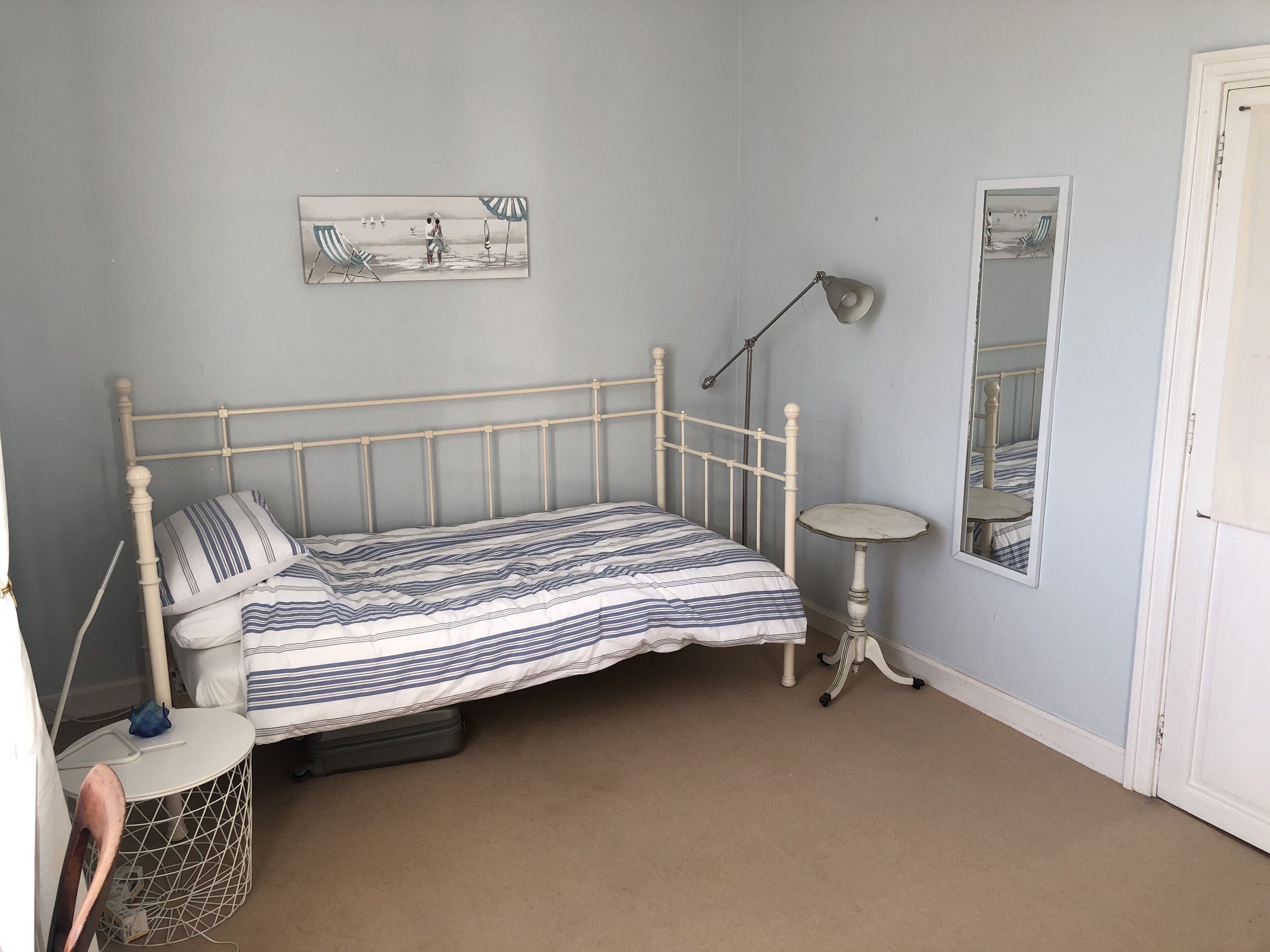 A single bed with a striped bedspread, a bedside table, a lamp, a small round table, a chair, and a wall-mounted picture.