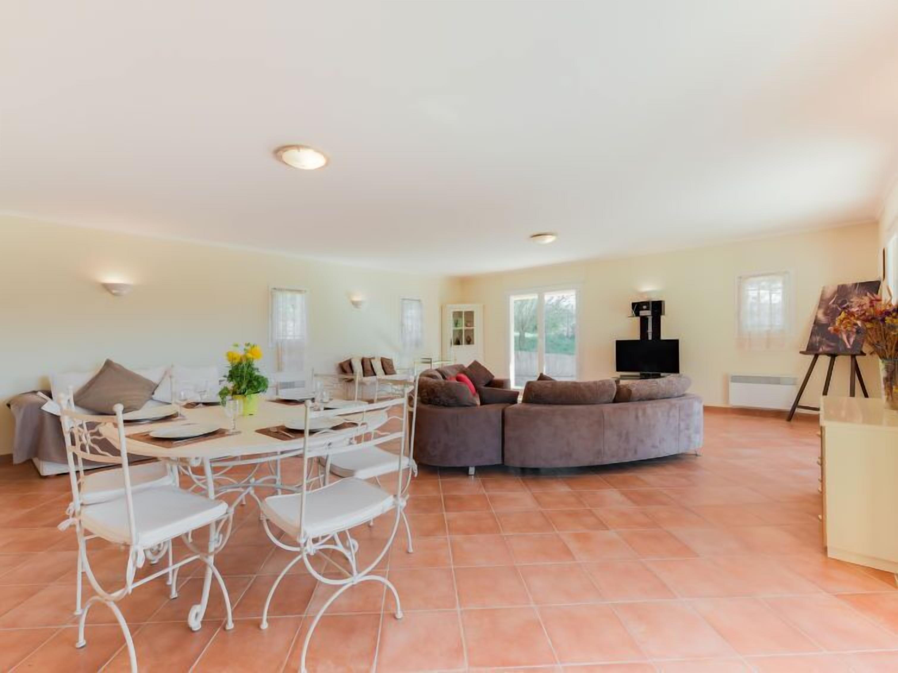 A spacious living room with a white metal dining table, white chairs, a grey sofa, and a TV.