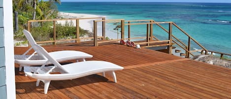 A wooden deck with white wicker lounge chairs overlooking a beach and ocean.