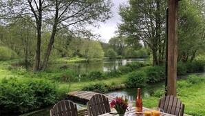A wooden table with chairs, a vase of flowers, a bottle, glasses, and a pitcher of orange juice, set against a backdrop of a river and greenery.