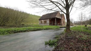 A rustic wooden house with a porch, a small stream running beside it, and a grassy hill in the background.
