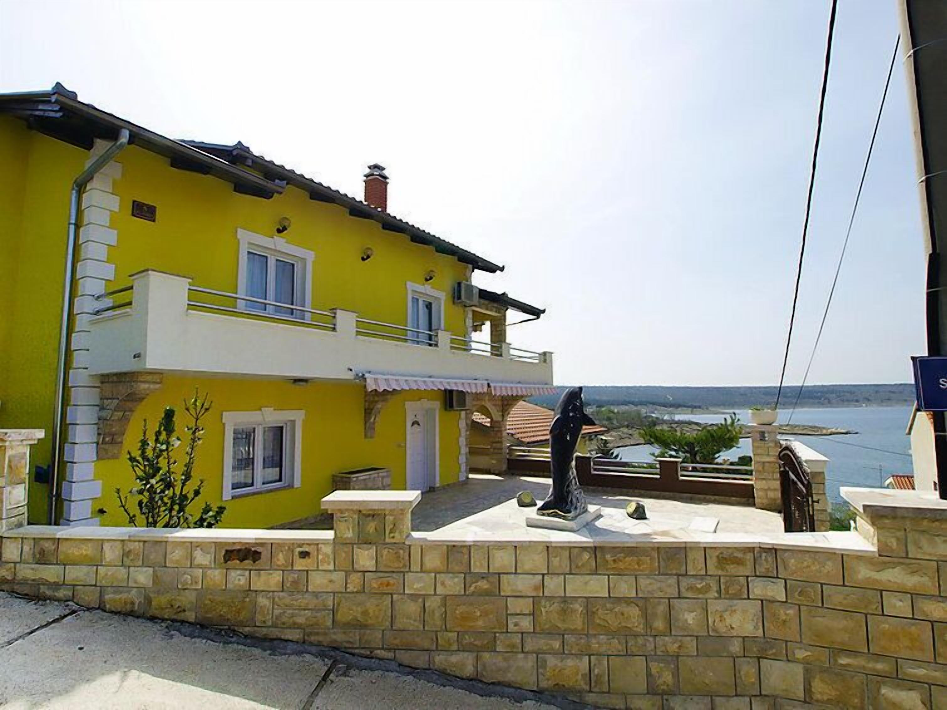 A yellow two-story house with a balcony, a stone wall, and a view of the sea.