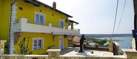 A yellow two-story house with a balcony, a stone wall, and a view of the sea.