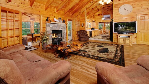 A wooden cabin interior with a fireplace, leather couches, a television, and a dining area.