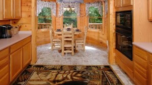 A wooden cabin interior with a dining area, kitchen, and windows with curtains.