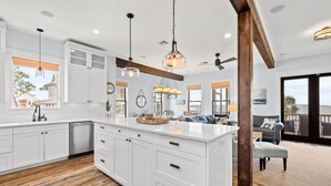 A spacious kitchen with white cabinetry, a large island, and wooden flooring. There is a dining area with a sofa and a view of the outdoors. - Baydream: Stunning Bayfront Home, Pet-Friendly, Hot Tub, Sleeps 12 (Port St. Joe)