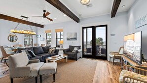 A spacious living room with a dark-colored sofa, a light-colored armchair, and a coffee table. There is a large window with a view of the outdoors, and a ceiling fan is visible. - Baydream: Stunning Bayfront Home, Pet-Friendly, Hot Tub, Sleeps 12 (Port St. Joe)