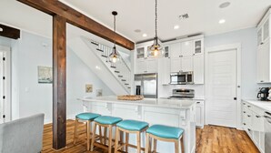 A modern kitchen with white cabinetry, a central island with bar stools, a wooden staircase, and a hanging light fixture. - Baydream: Stunning Bayfront Home, Pet-Friendly, Hot Tub, Sleeps 12 (Port St. Joe)