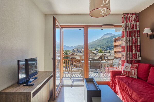 Habitación con televisión sobre un mueble de madera, sofá rojo, puerta corrediza de cristal que da a una terraza exterior con mesas y sillas, y vistas a la montaña.