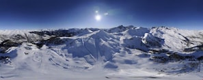 Vista panorámica de un paisaje montañoso cubierto de nieve, con cielo despejado y sol brillante.