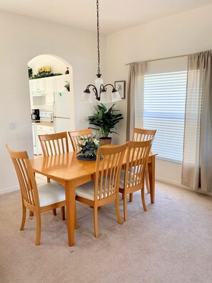 Un comedor con una mesa y sillas de madera, una lámpara colgante, una planta en maceta y una cocina al fondo.