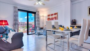Una sala de estar moderna con sofá, mesa de comedor y sillas. Se ve una ventana grande con vistas a edificios y un paisaje urbano.