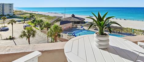 A balcony with a view of a beach, a pool, and a building in the background.