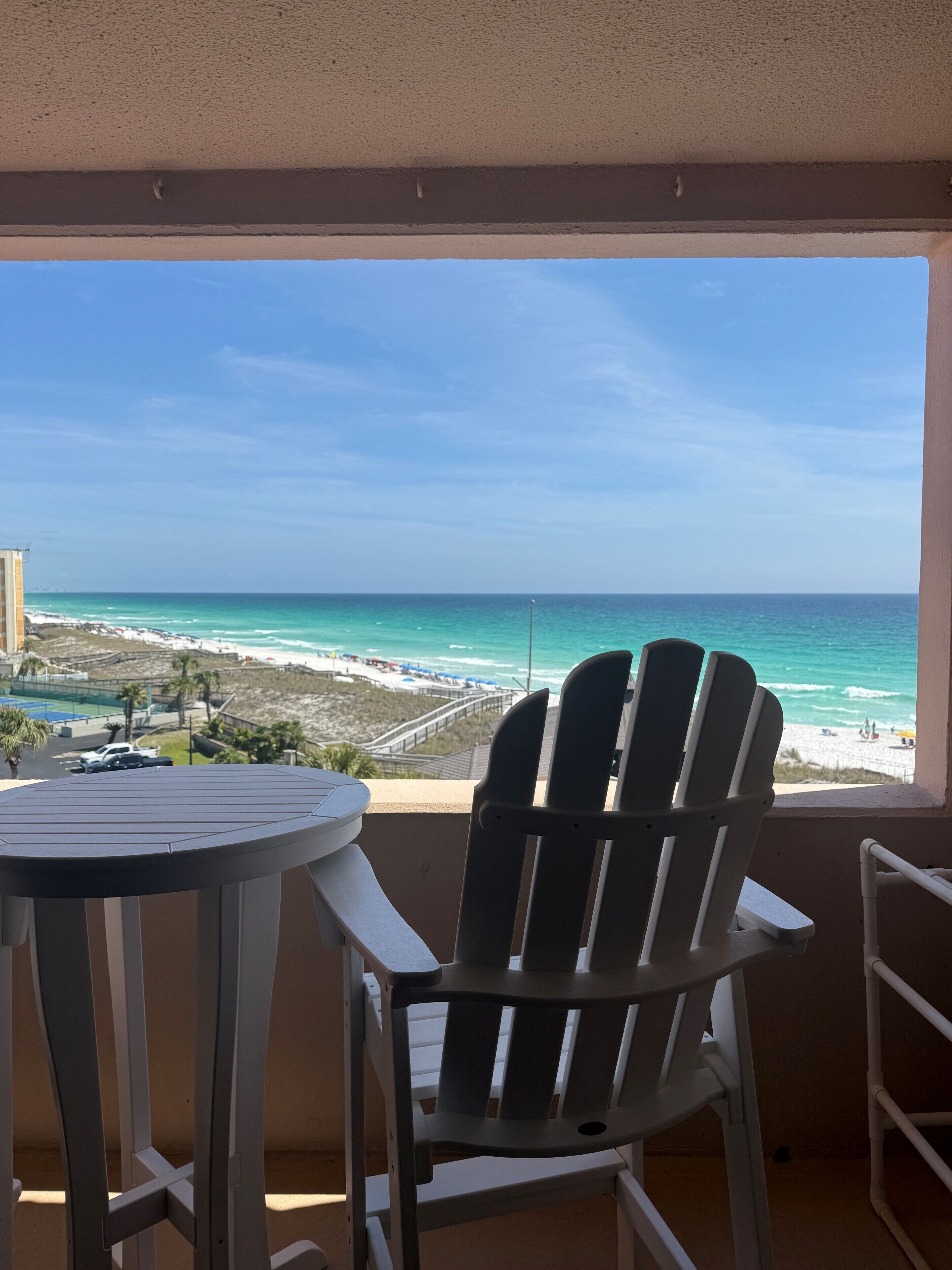 A balcony with a view of the beach, a chair, and a table.