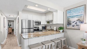 A modern kitchen with white cabinets, stainless steel appliances, granite countertops, and a painting of a beach scene. - Gulf view unit at Jetty East! One free beach set up daily. (Destin)