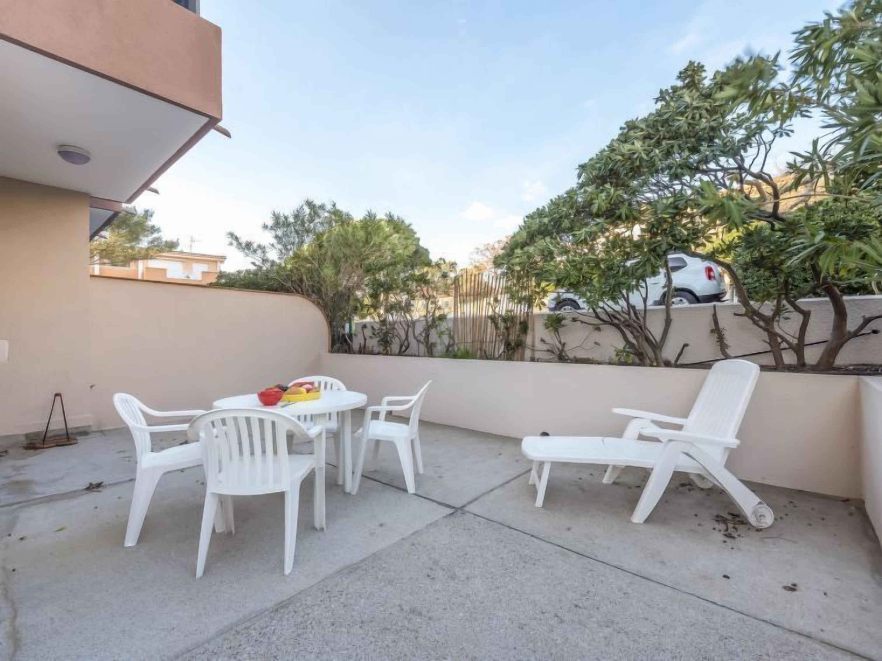 Un patio al aire libre con sillas y mesa de plástico blanco, rodeado por un muro bajo y follaje.