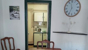 Una sala da pranzo con un tavolo rosso, quattro sedie di legno, una sveglia, un quadro appeso al muro e una cucina sullo sfondo.