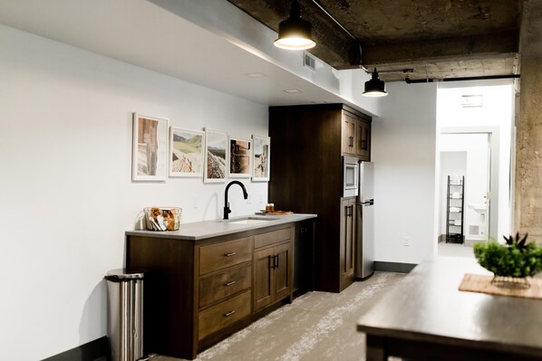 A modern kitchen with dark wooden cabinets, a stainless steel sink, and a countertop with a basket of bread. There are framed pictures on the wall and a plant on the counter.