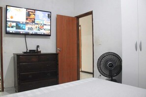 A bedroom with a television mounted on the wall, a wooden wardrobe, a bed with a white bedspread, and a standing fan.