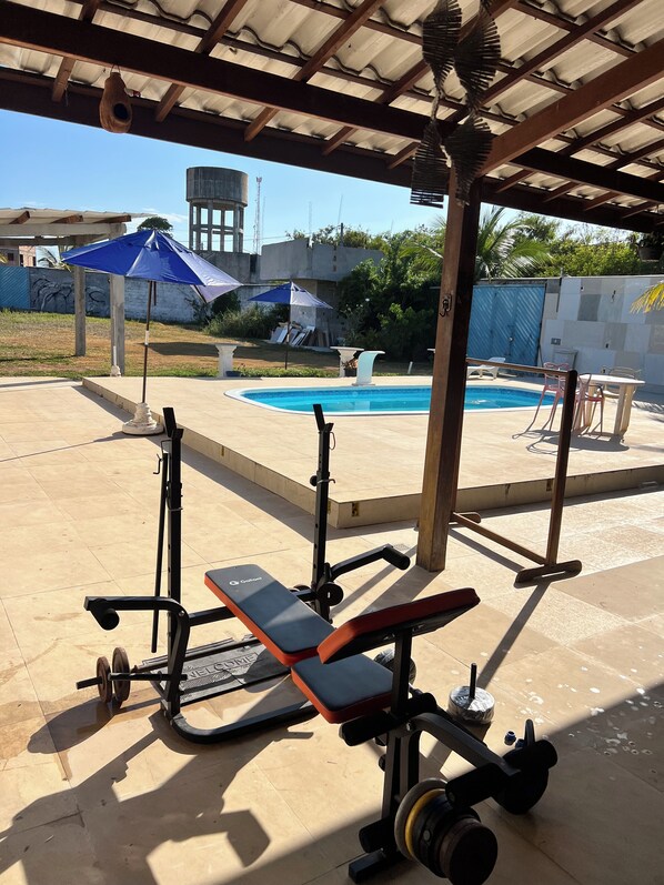 A covered outdoor area with a pool, exercise equipment, and a patio with chairs and umbrellas.