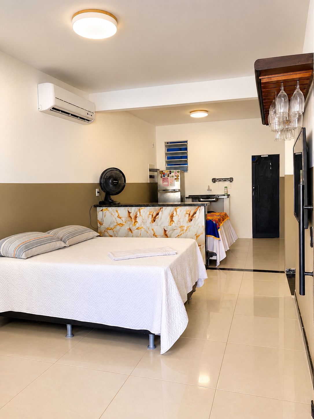 Quarto de hotel com cama, escrivaninha com computador, ar condicionado e ventilador.