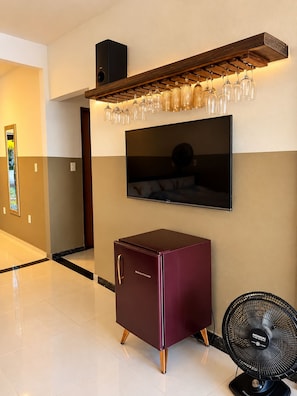 Habitació d'hotel moderna amb televisió de pantalla plana penjada a la paret, nevera petita, ventilador de peu i una prestatgeria de vi amb gots.