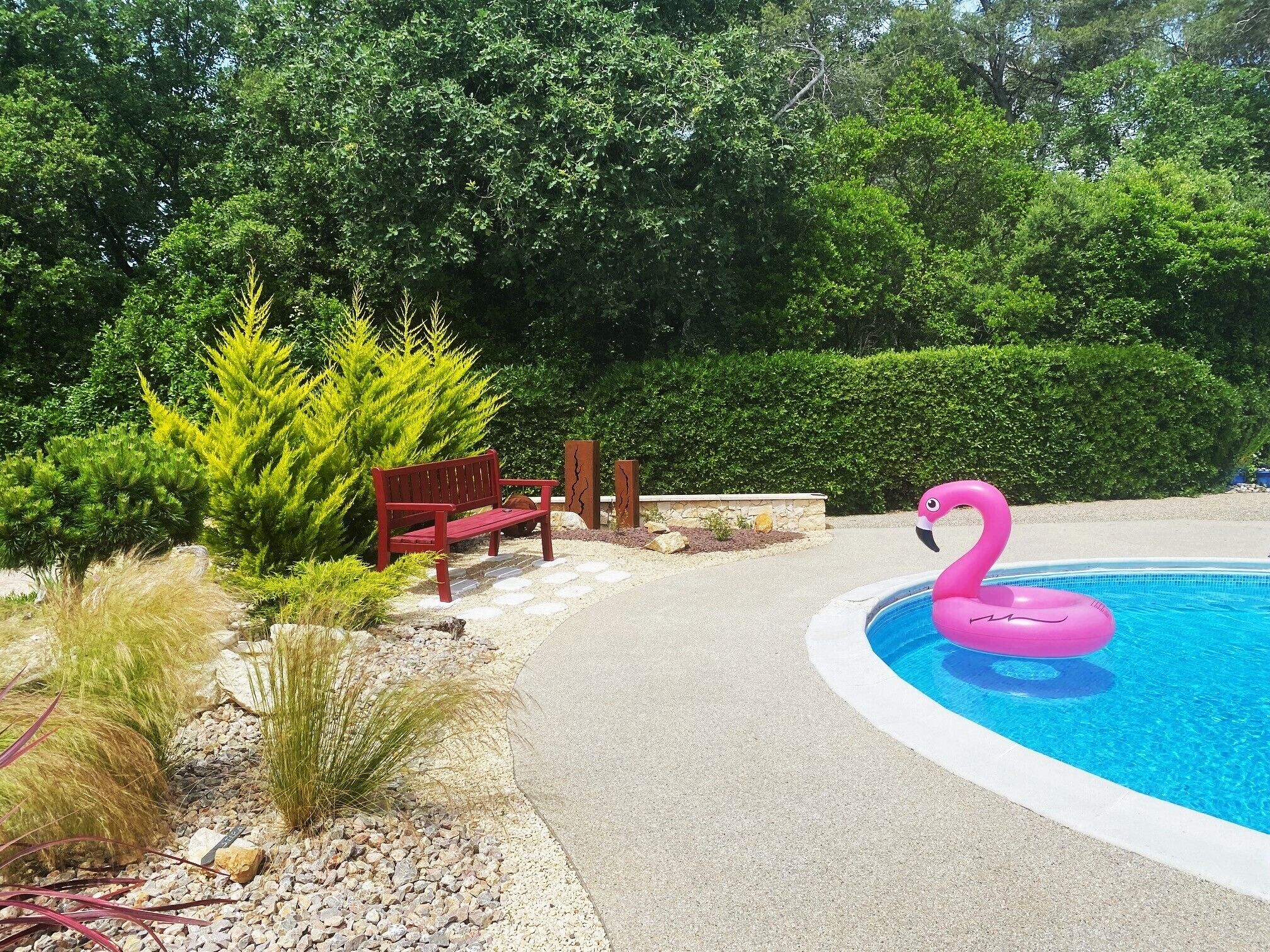 Une piscine agrémentée d’un flotteur en forme de flamant rose, entourée d’un chemin, d’un banc et de diverses plantes.